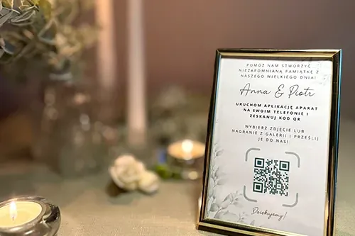 Kartka z kodem QR w złotej ramce na weselnym stole przy świecach.